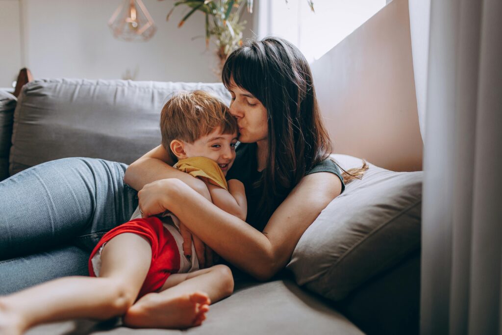 A tender moment of a mother kissing and hugging her child on a cozy sofa indoors.
