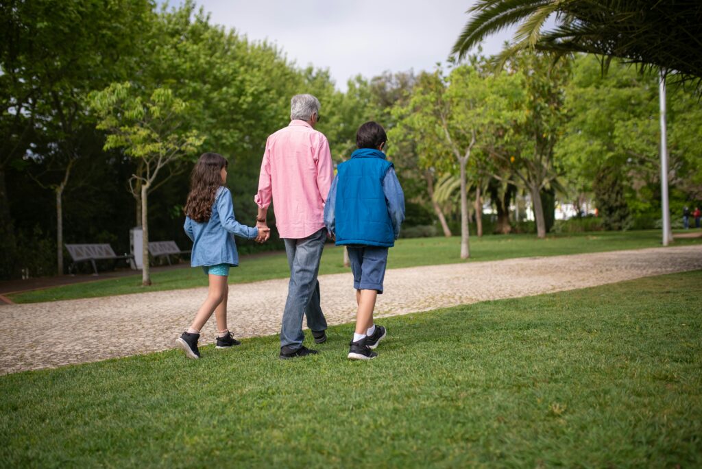 A grandfather holds hands with his grandchildren on a relaxing walk in a lush green park.
